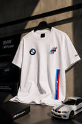 A white BMW Motorsport t-shirt hanging on a black hanger against a dark gray wall. The shirt features the BMW roundel logo on the left chest, a M logo graphic on the right chest, and a vertical blue, white, and red M stripe on the right sleeve.