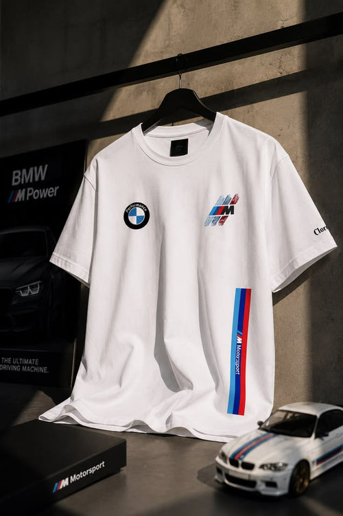 A white BMW Motorsport t-shirt hanging on a black hanger against a dark gray wall. The shirt features the BMW roundel logo on the left chest, a M logo graphic on the right chest, and a vertical blue, white, and red M stripe on the right sleeve.